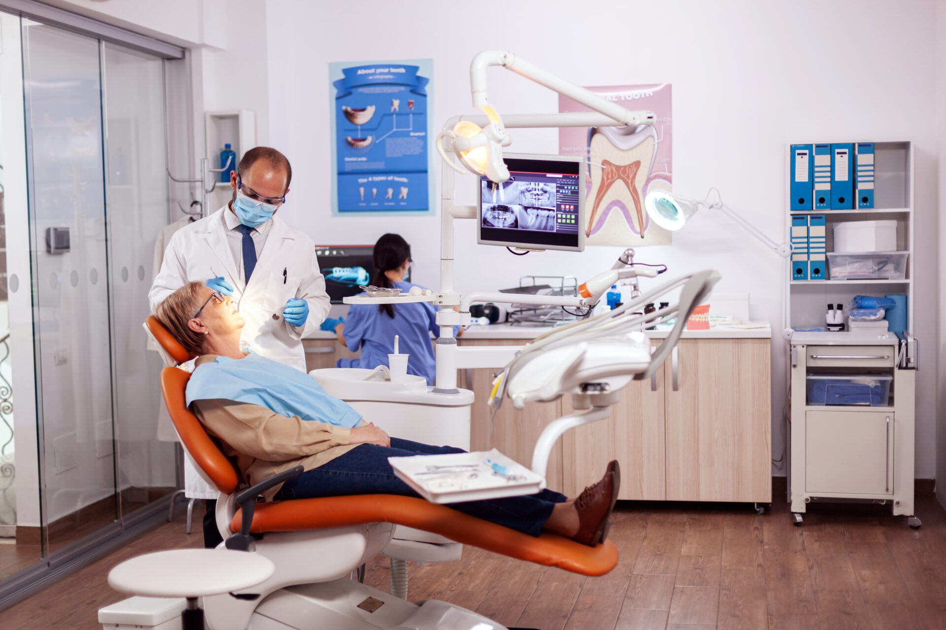 Dentist doing a dental treatment on a elderly patient. His assistant is looking at the screen with teeth x-ray.