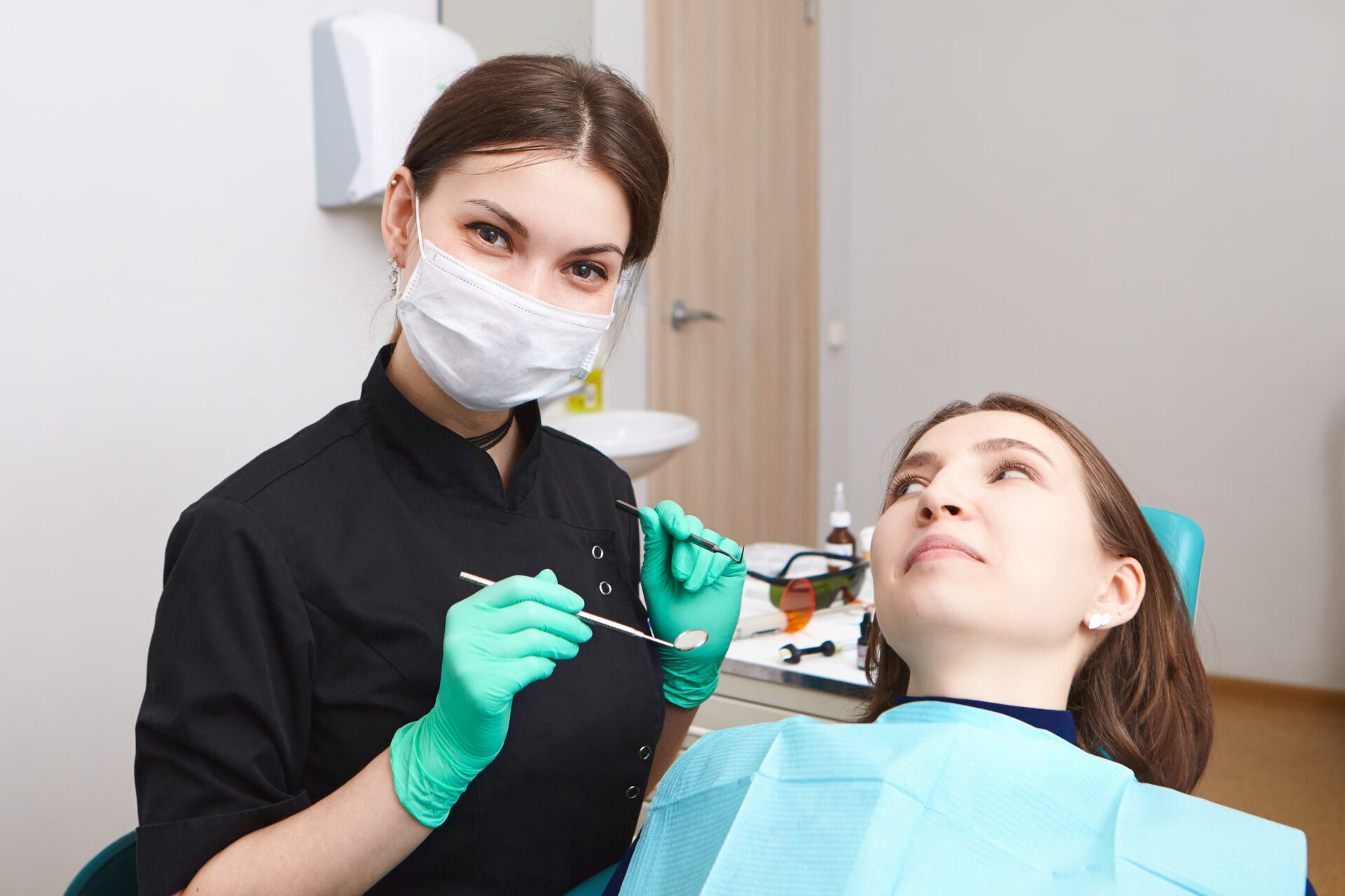 Indoor shot of skillful confident young female dentist wearing exam gloves and white mask holding metal probe and dental miror, ready to examine oral cavity of woman patient sitting in chair